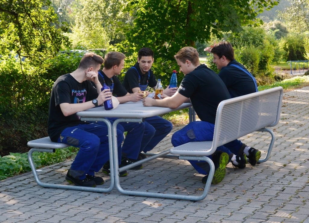 Produktionsmitarbeiter im Blaumann sitzen draußen auf einer Sitz-Tisch-Kombination Parador und machen Pause